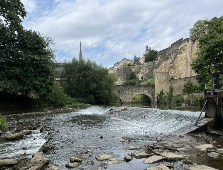 KeyInn Serviced Residences Un pont de pierre et de hauts murs de forteresse surplombent une rivière douce avec un petit barrage, entourée d'arbres luxuriants. À l'arrière-plan, des bâtiments historiques et une flèche d'église s'élèvent au-dessus des falaises sous un ciel partiellement nuageux.