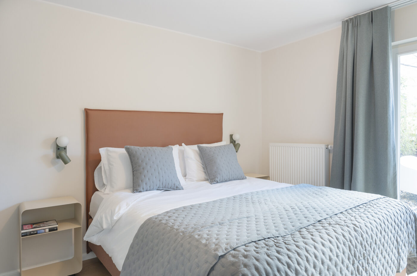 Keyinn Serviced Residences Belair bedroom with a double bed, tan headboard, and gray pillows. The space features a side table with books, soft gray window curtains, and a clean minimalist design.