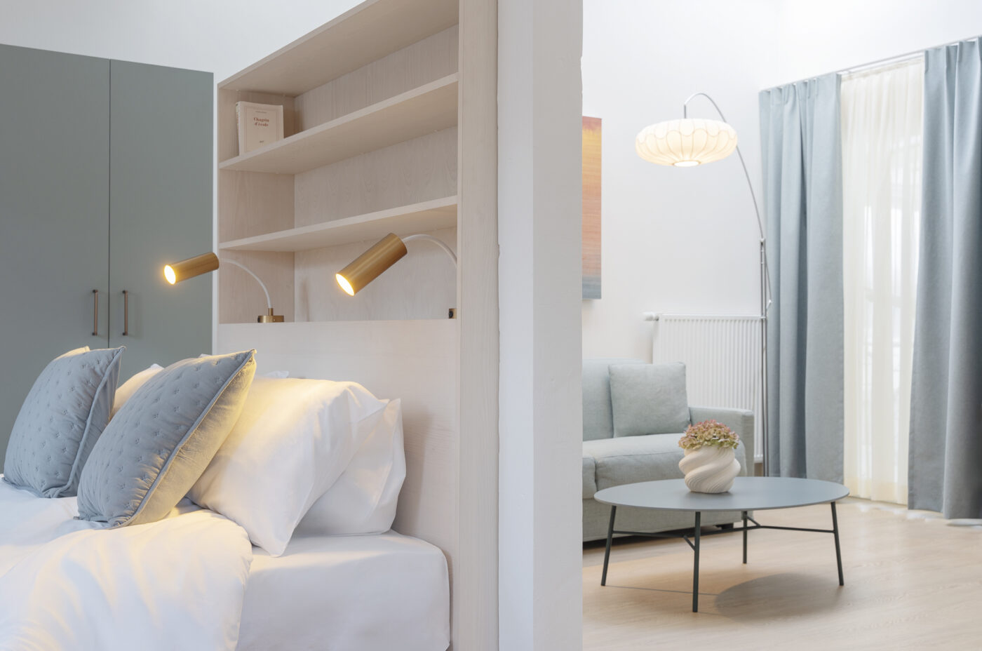 Keyinn Serviced Residences Belair bedroom with white bedding, blue pillows, and shelf-mounted lamps. The space includes a background living area with a sofa, table, and soft lighting.