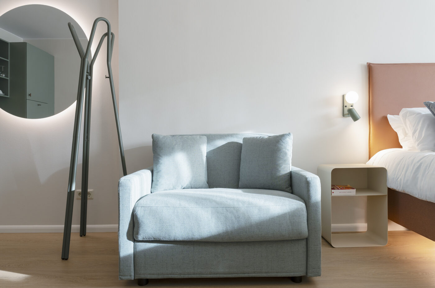 KeyInn Serviced Residences Une chambre d'hôtel moderne et minimaliste avec un fauteuil bleu clair, un miroir mural rond, un porte-manteau sur pied, une table de chevet avec un livre, un lit avec des draps blancs et une lampe murale.