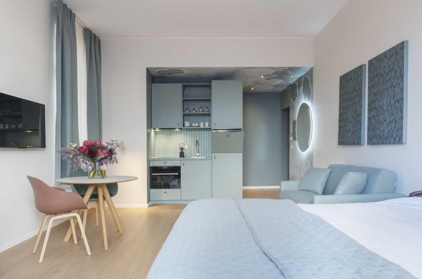 Keyinn Serviced Residences Belair studio with a pastel blue theme, kitchenette, and sofa. The space includes a dining table, flowers, soft lighting, and abstract art.