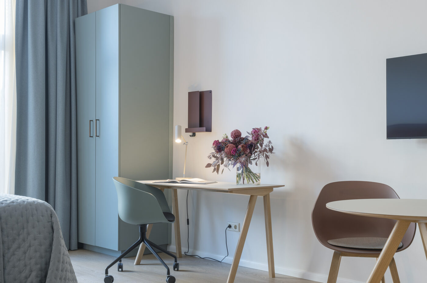 Keyinn Serviced Belair Residences room with a light blue wardrobe, wooden desk, and lamp. The space includes a teal chair, round table, and floral accents.