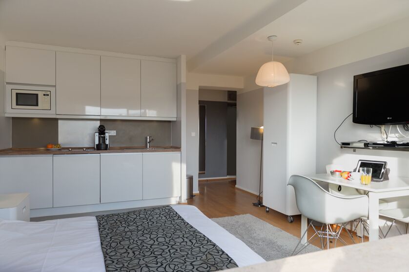 A polished Keyinn Serviced Residences studio apartment featuring a streamlined kitchen with white cabinets, a single bed, and a dining table with chairs, and a wall-mounted TV.