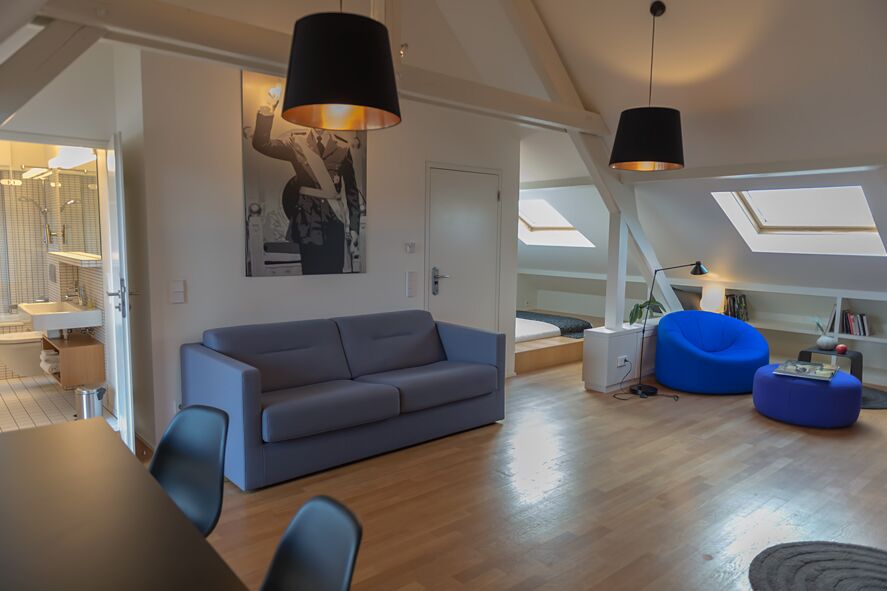 Keyinn Serviced Residences Parc de Merl loft with grey and blue seating, black pendant lighting, and sun-drenched skylights.