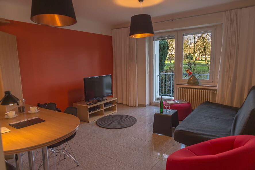Keyinn Serviced Residences Parc de Merl living room with a red wall, round dining set, featuring Large windows & balcony door overlooking the park.