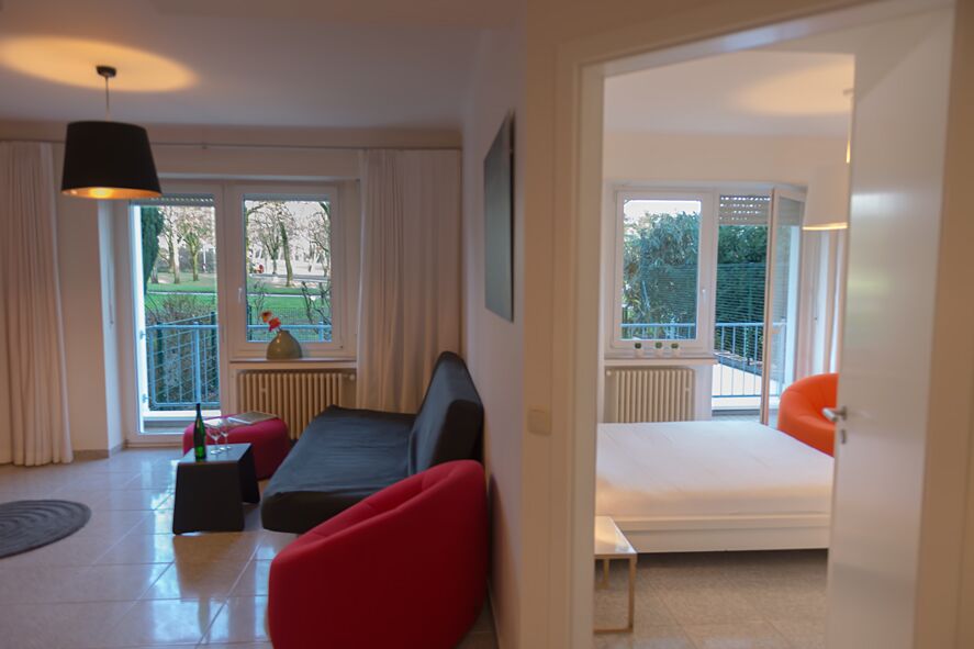 Keyinn Serviced Residences Parc de Merl space with a black sofa, bold red seating, and a dining set overlooking the park. The sleek transition to the white-toned bedroom offers a clear, well-appointed, and tranquil atmosphere.