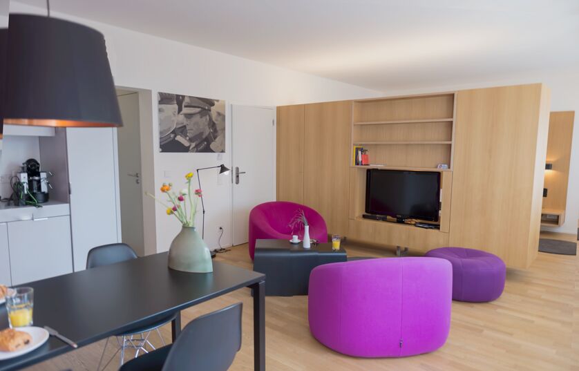 Keyinn Serviced Residences Parc de Merl living space with vibrant pink seating, a purple ottoman, and wooden flooring. With a black dining table and wooden TV unit.