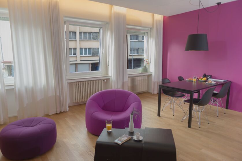 Keyinn Serviced Residences Parc de Merl room with a pink wall, pink chairs, and wooden flooring. Large windows and white curtains are included.
