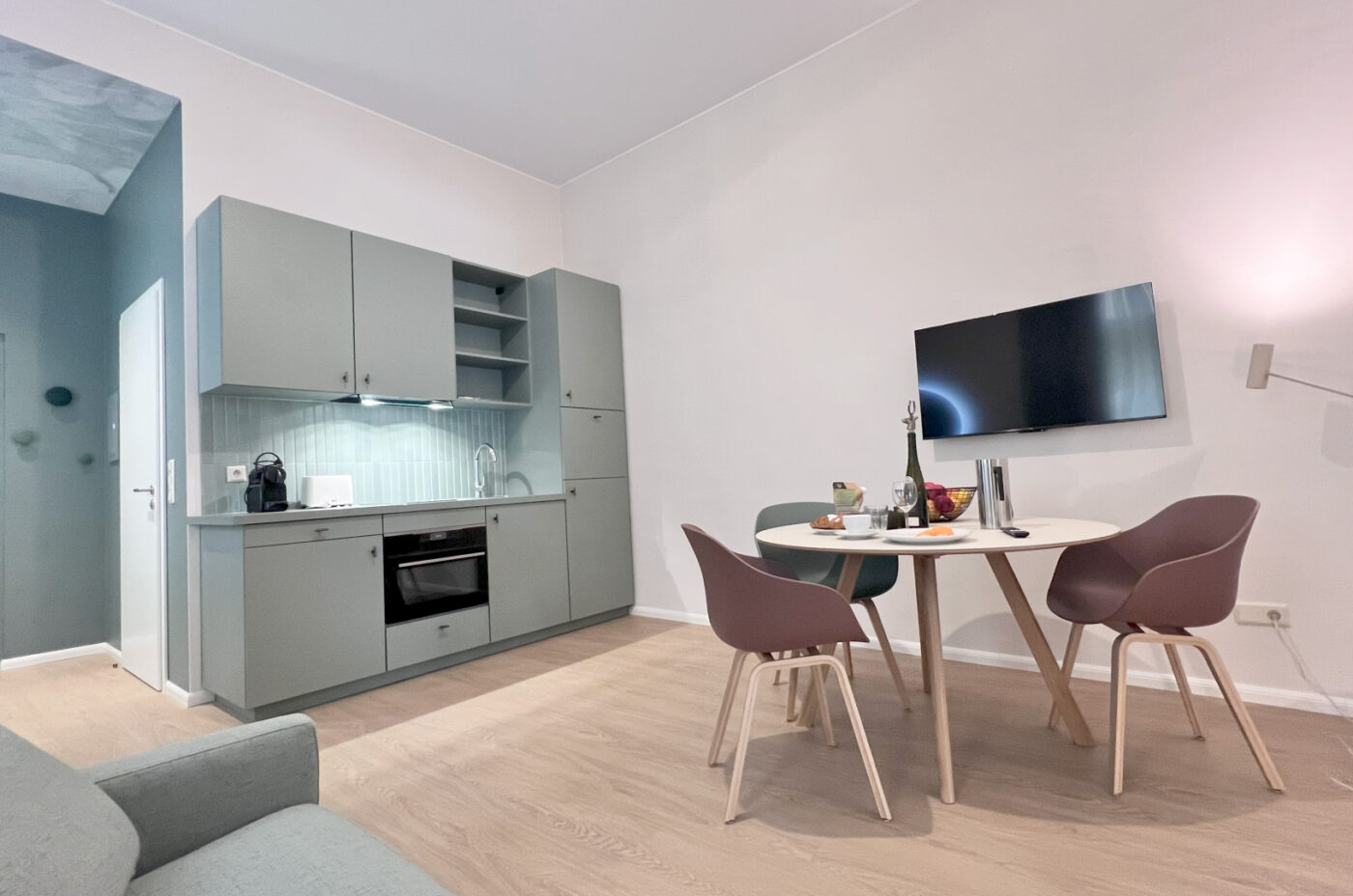 Keyinn Serviced Residences Belair kitchen with light gray cabinets and a built-in oven. The space features a round dining table for two set under a wall-mounted TV with light wood floors throughout.