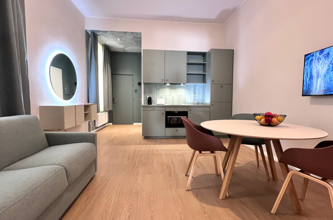 Keyinn Serviced Residences Belair living room with light wood floors, a gray sofa, and a wall-mounted TV. The space includes a round dining table with four chairs and a kitchen in the background.