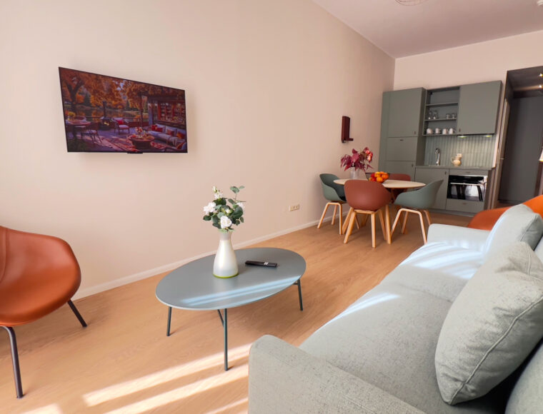 Keyinn Serviced Residences apartment living room featuring a light gray sofa and a bold orange armchair on wooden flooring. A floral centrepiece rests on a gray coffee table, with a glimpse of the kitchen in the background.