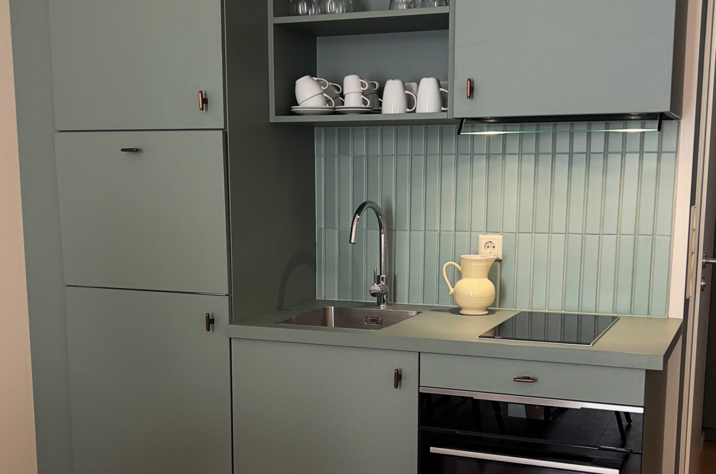 Keyinn Serviced Residences Belair kitchen with pastel green cabinets, a stovetop, and an open shelf with white mugs. The space features a yellow pitcher on the counter and a clean, modern design.