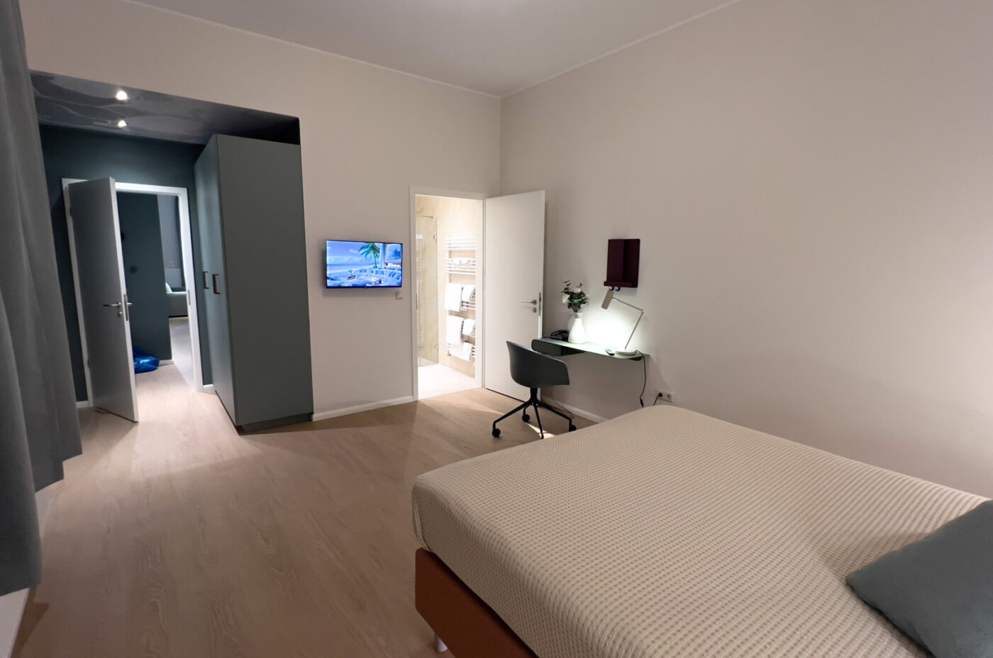 Keyinn Serviced Residences Belair bedroom with neutral tones, a desk, and a chair. The space features a wall-mounted TV and an open view into a bright, modern bathroom.