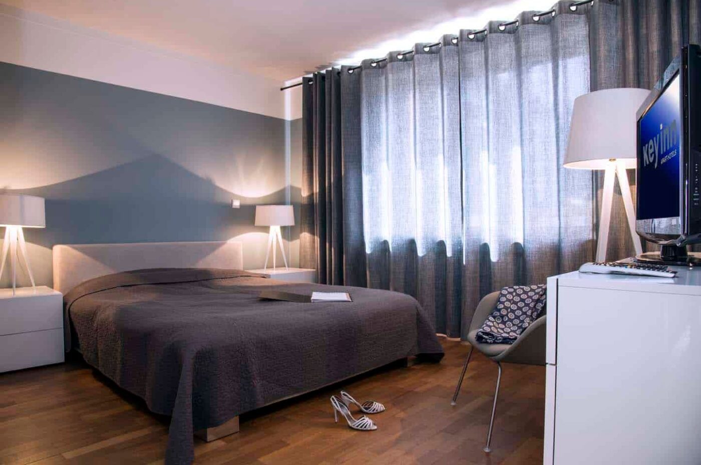 A bedroom at Keyinn Serviced Residences featuring a gray bedsheets and curtains, with blue themed wall. White lamps and a TV positioned on a dresser.