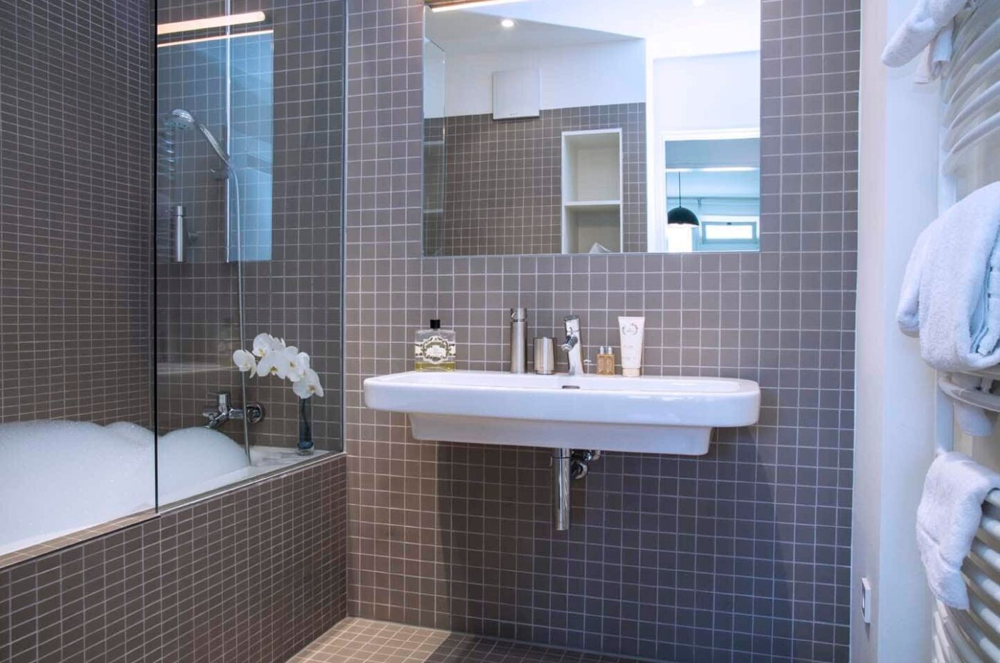 Keyinn Serviced Residences Limpertsberg bathroom with grey tiling, a sleek sink, and a bathtub featuring a glass partition and bubble bath.