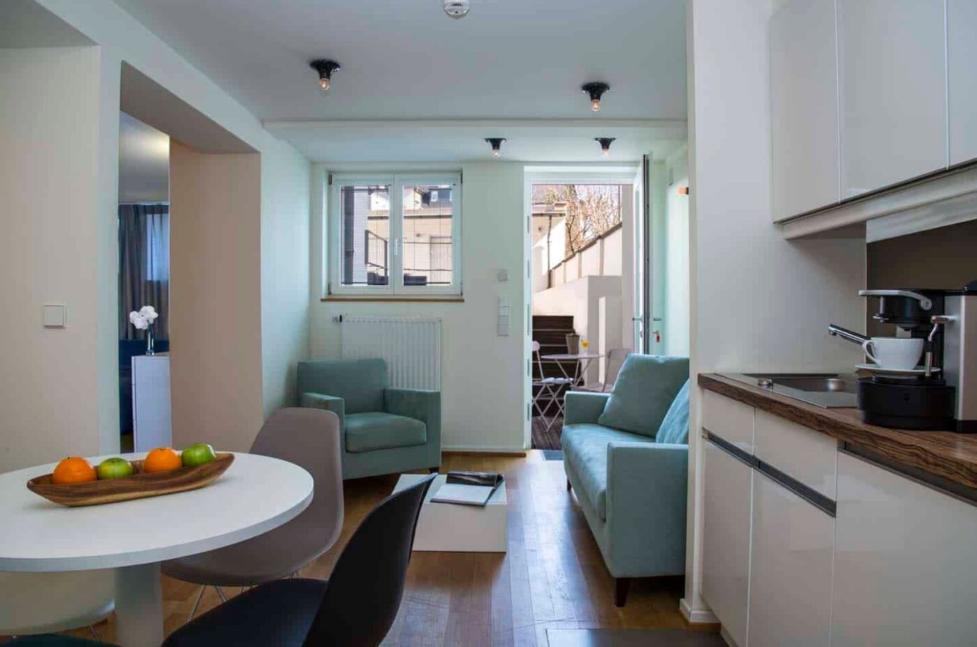 Keyinn Serviced Residences Limpertsberg flat, with a sleek kitchenette, a round dining set, and a green sofa. The layout offers a view of the patio.