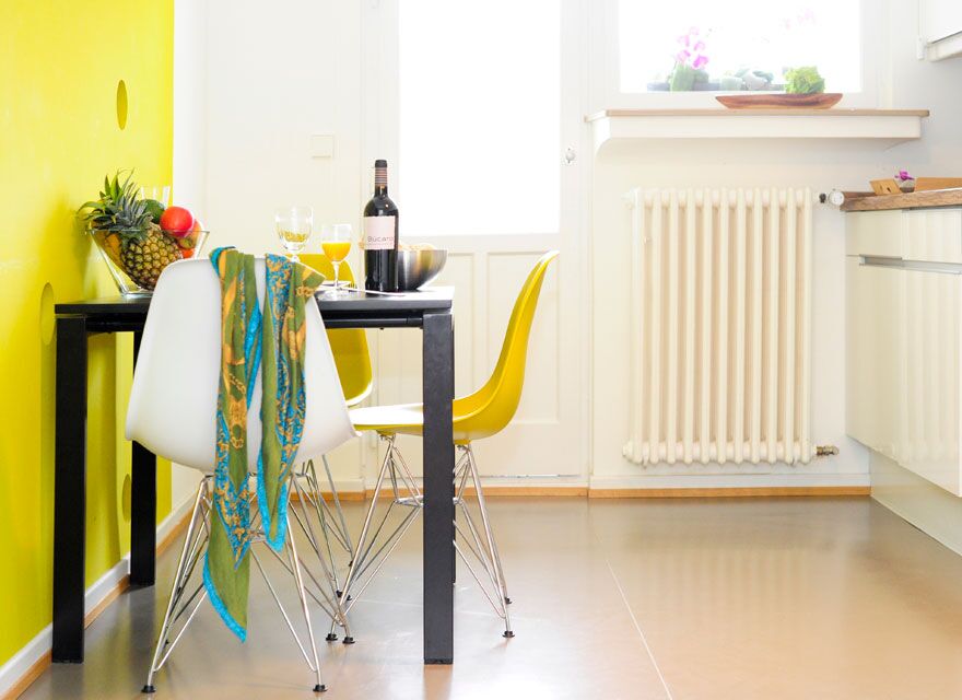 Bright, modern kitchen at Keyinn Serviced Residences in Limpertsberg. with a two-person black dining table with white and yellow chairs, yellow wall, fruit, wine & a patterned scarf.