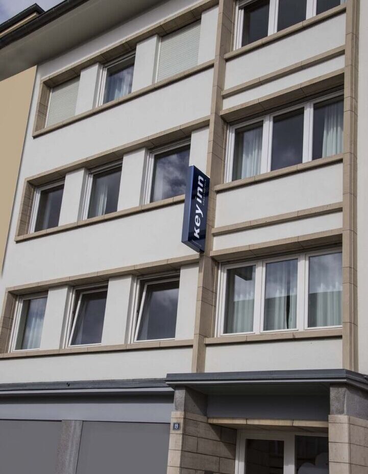 The image shows the Keyinn Serviced Residences building in Parc de Merl.. With a guest entrance, and four floors of windows.