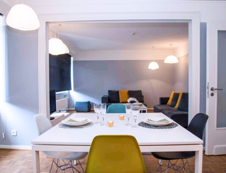 KeyInn Serviced Residences A modern dining and living room with a white table set for four, surrounded by mixed chairs. In the background, gray sofas with yellow cushions, pendant lights, and large windows with black blinds.