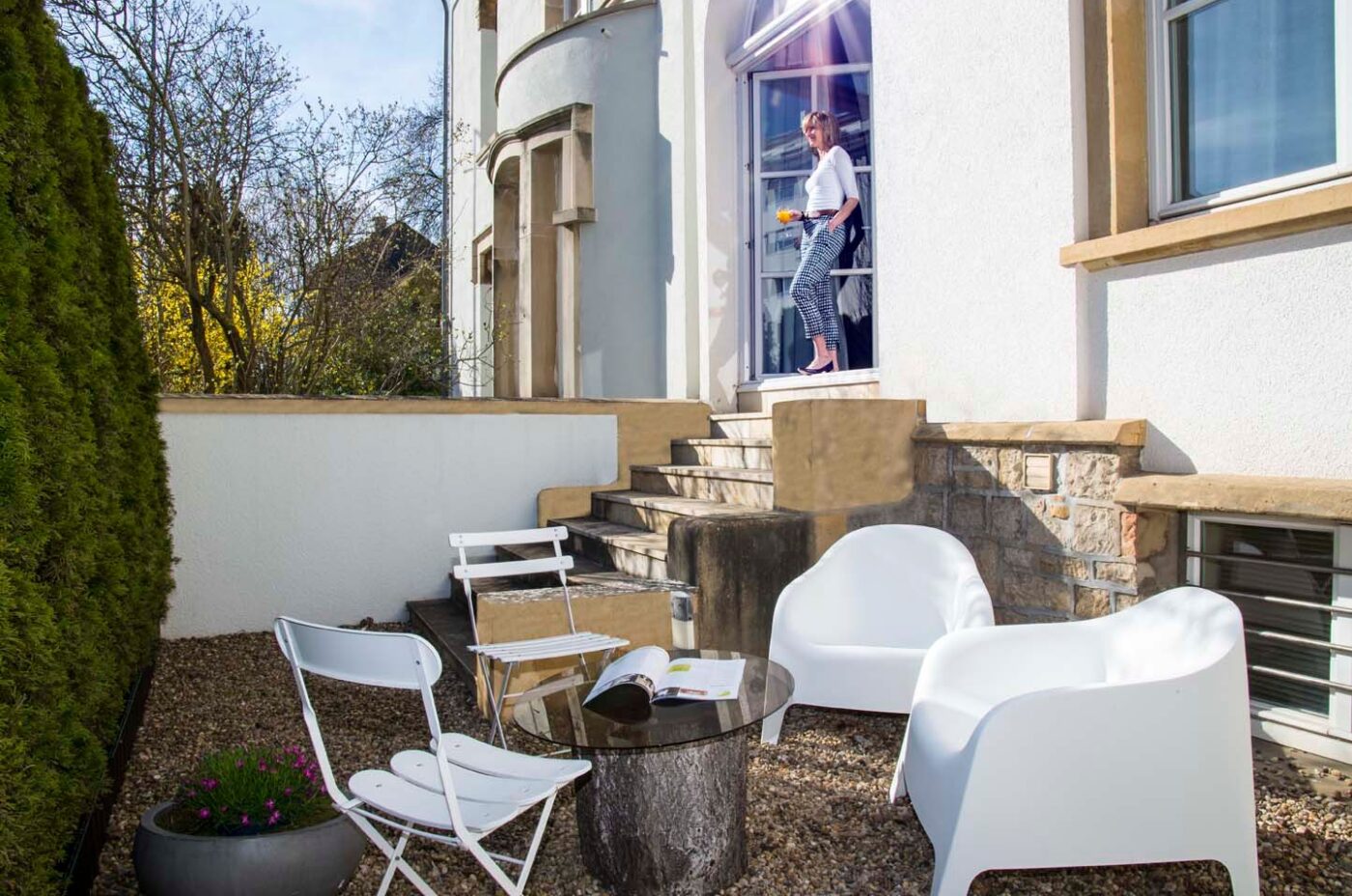 Sunlit patio with modern white chairs and a glass-top table on gravel at Keyinn Serviced Residences. A person stands on stairs holding a tray near an arched doorway.