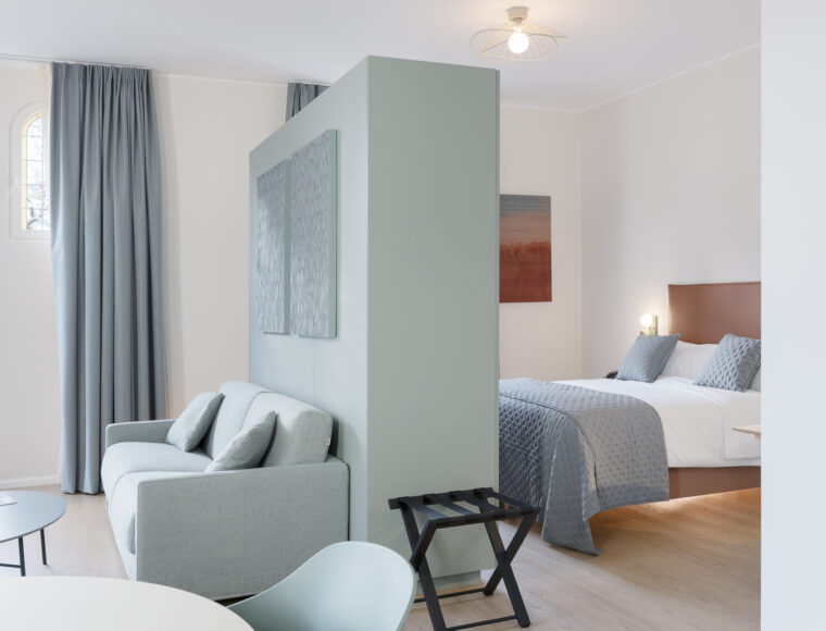 KeyInn Serviced Residences Modern studio apartment with a soft blue and gray color scheme, featuring a bed and sofa separated by a partial wall, large windows with curtains, and minimalist decor.
