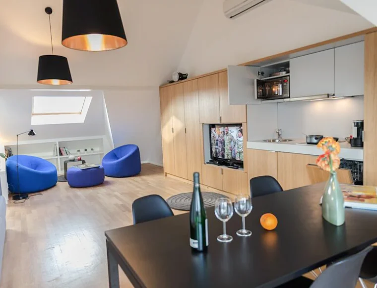 Open-plan apartment at Key-inn Serviced Residences Parc de Merl with wooden floors, integrated kitchen, dining table set for two, TV, and two blue chairs.