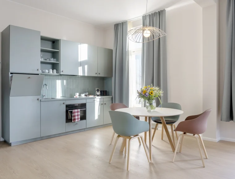 KeyInn Serviced Residences Modern kitchen with light blue cabinets, built-in oven, and a dining area featuring a round table with four colorful chairs. A bouquet of flowers sits on the table; large windows let in natural light.